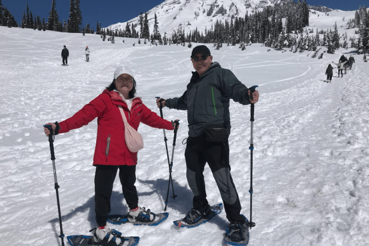 Two people snowshoeing on a snowy mountain slope with clear blue sky.