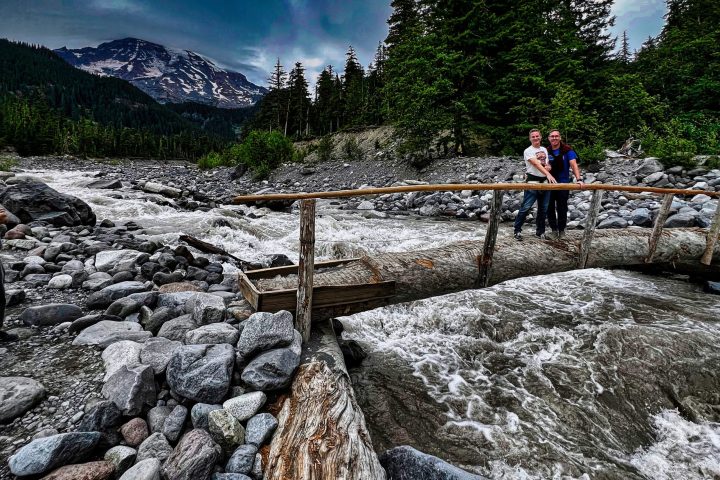 two people standing on a wood bridge over a river with mt rainier behind them on a day trip to mt rainier from seattle