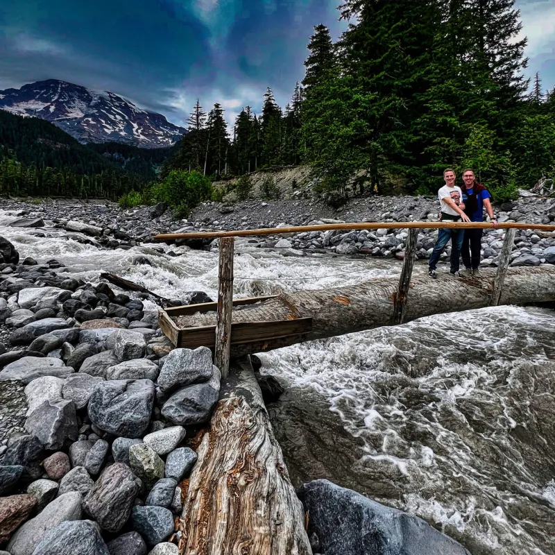 two people standing on a wood bridge over a river with mt rainier behind them on a day trip to mt rainier from seattle
