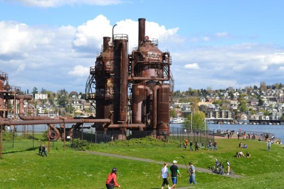 Park scene with industrial gasworks structure and people walking, biking, and relaxing on a sunny day.