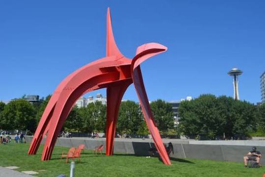 Red abstract sculpture in a park with trees and city skyline.