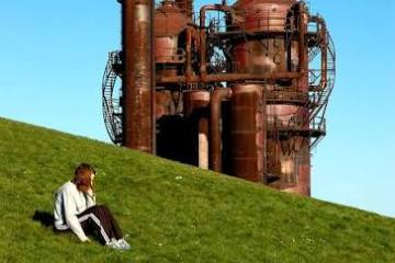 Person sitting on grassy hill with rusted industrial structure in background.
