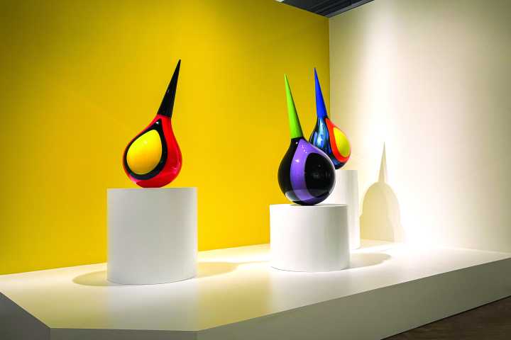 Colorful glass sculptures on pedestals against a yellow wall in a gallery.