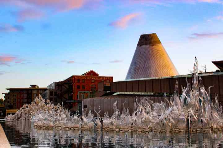 Glassy sculptures and cone-shaped building at sunset by reflective water.