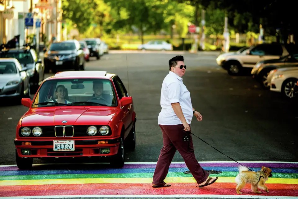 Person walking a dog across a rainbow crosswalk in front of a red car.