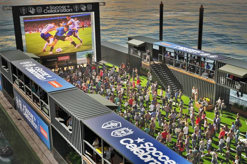 Crowd watching soccer match on outdoor screen at waterfront venue.