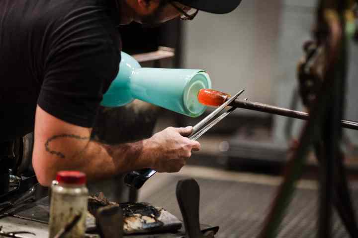 Man shaping molten glass on a rod with tongs, creating a blue glass object.