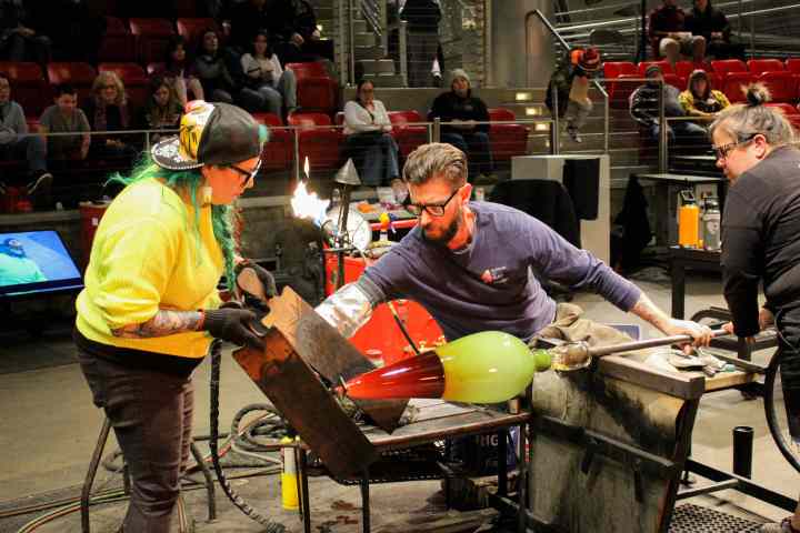 Glassblowers shaping a glass piece with glowing tools in a workshop, surrounded by spectators.