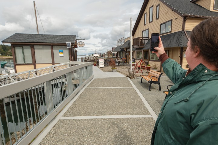 Person in green jacket takes photo of waterfront shops and boats on a cloudy day.