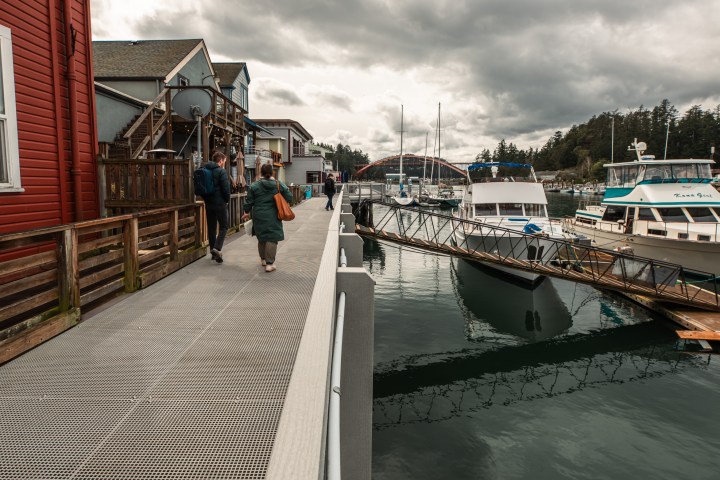People walking on a dock beside boats and waterfront buildings under cloudy skies.