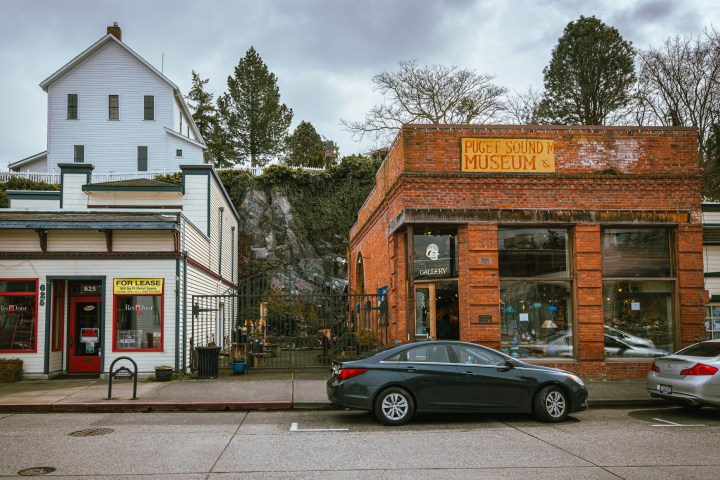Street view of small shops and a museum in brick and wood buildings with cars parked in front.