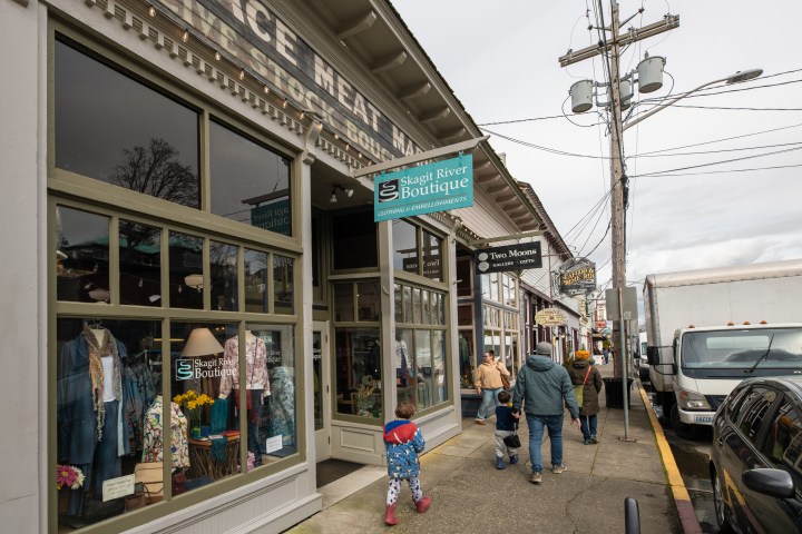 People walking by Skagit River Boutique on a street lined with shops and parked cars.