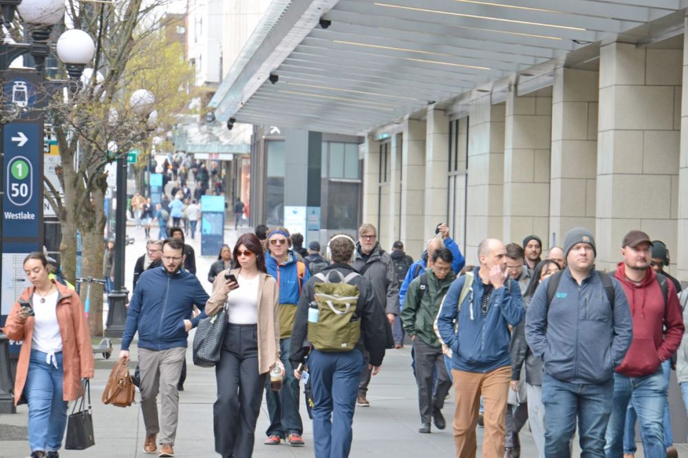 Crowd of people walking on city sidewalk near buildings, some using phones.
