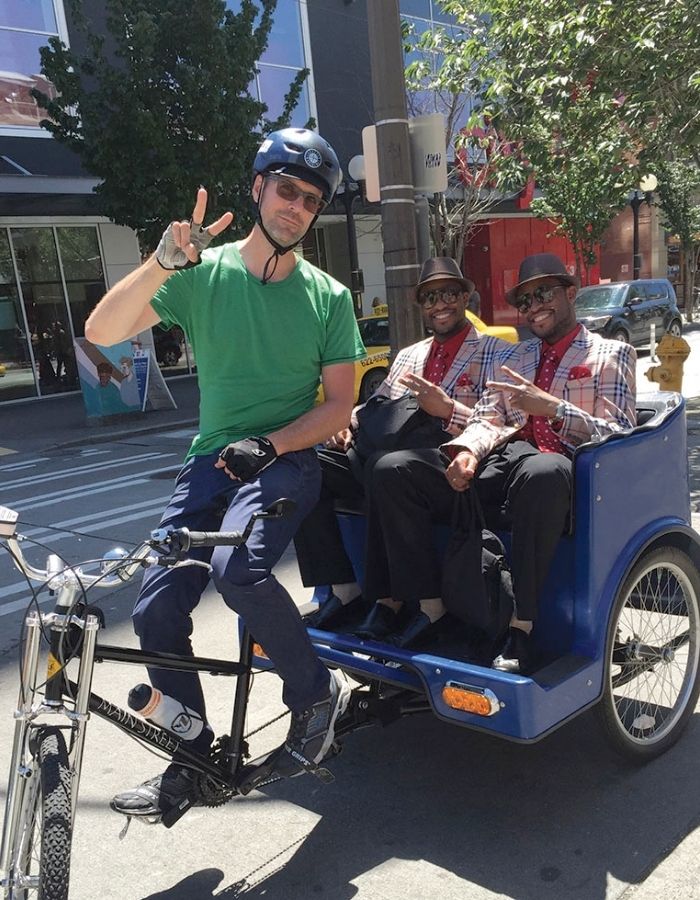 Man in helmet on rickshaw bike with two passengers wearing hats and sunglasses.