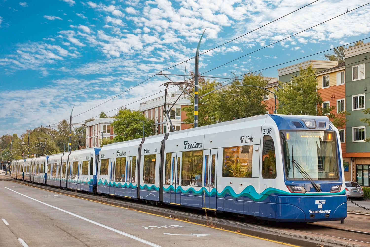 Light rail train on urban tracks with trees and buildings in background.