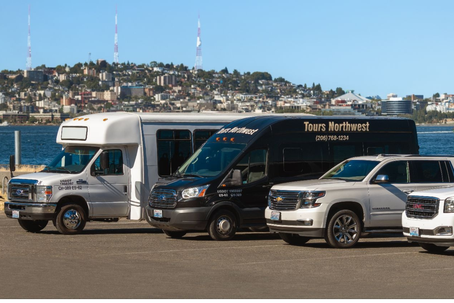 Four tour vehicles parked by a waterfront with cityscape in the background.