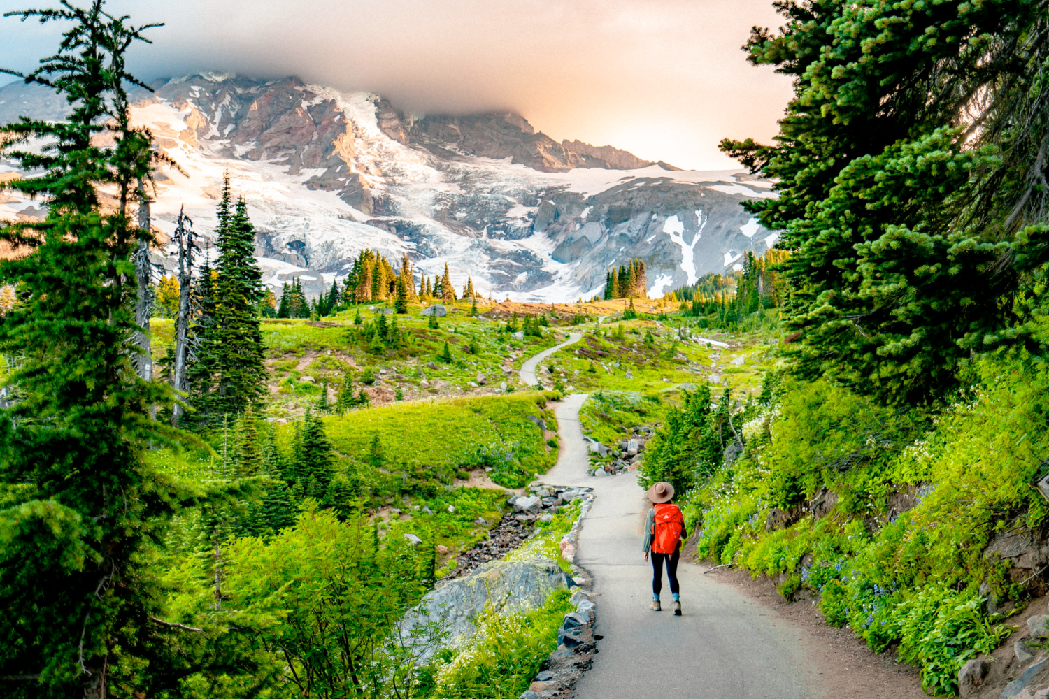 Person with backpack walks on trail towards snow-capped mountain under cloudy sky.