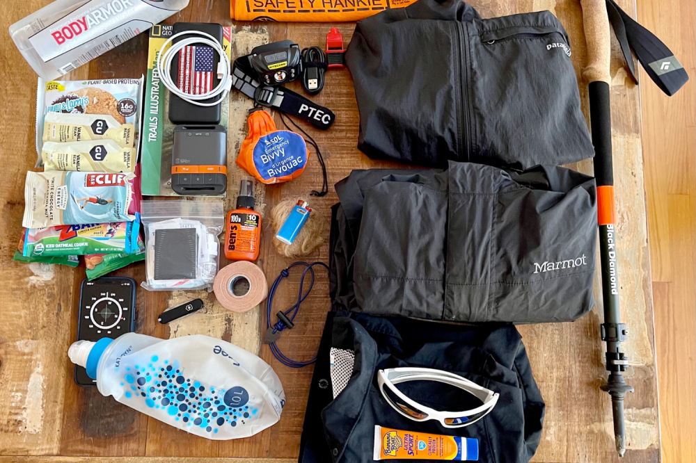 The 10 Hiking Essentials Assorted outdoor gear including jackets, climbing tools, snacks, sunscreen, and hydration packs on a table.